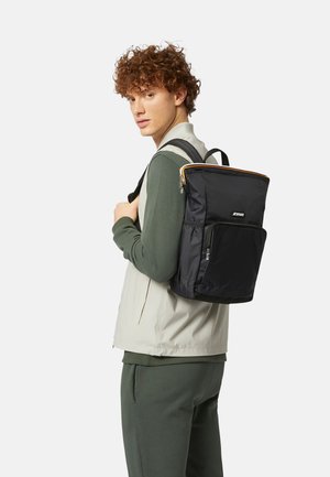 Giovane uomo con capelli ricci che indossa abiti verdi e beige, con uno zaino nero con più scomparti con cerniera.