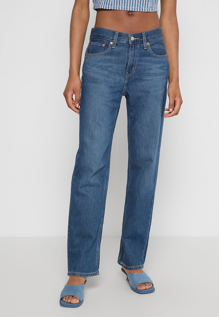Helle blaue Jeans mit geradem Bein, Fünf-Taschen-Design und sichtbaren Nähten. Kombiniert mit passenden blauen Slidesandalen.