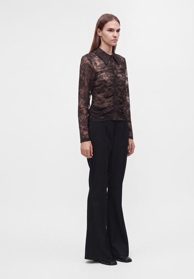Femme debout portant un chemisier en dentelle noire transparente à manches longues avec col et un pantalon noir à jambes larges sur un fond uni.