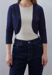 Granatowy krótki kardigan nałożony na beżowy prążkowany top, w połączeniu z wysoko w pasie ciemnymi dżinsami. Gładka faktura i dopasowany krój.