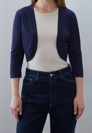 Cardigan court bleu marine sur un haut beige en rib, associé à un jean en denim foncé taille haute. Texture lisse et design ajusté.