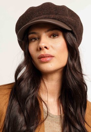 Woman with long dark hair wearing a brown wool cap with a leather brim, beige knit top, and camel-colored coat against a plain background.