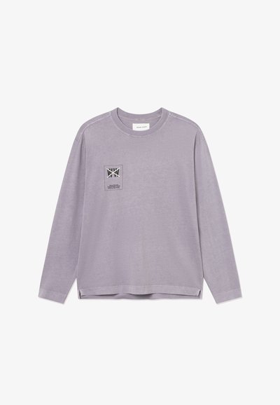 Chemise lavande à manches longues avec encolure ronde et petite étiquette logo imprimée sur le côté gauche de la poitrine, présentant des éléments de texte et graphiques.