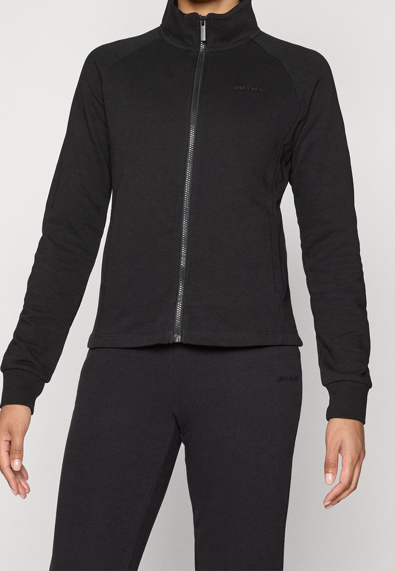 Chaqueta negra con cremallera y cuello alto, hecha de un tejido suave. Con mangas largas y una silueta ajustada. Branding mínimo en el pecho.