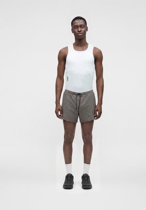 Jonge man staat, draagt een wit mouwloos shirt, grijze korte broek, witte sokken en zwarte sneakers tegen een effen lichte achtergrond.