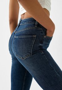 Jeans ajustados de denim en azul oscuro con una textura suave, que presentan una cintura de altura media, dos bolsillos traseros y costuras en contraste.