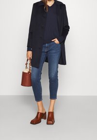 Námořnický kabát, tmavý svetr s výstřihem do V, modré úzké džíny, hnědé kožené boty na podpatku a béžová taška typu bucket s pleteným uchem.