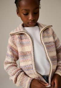 Strickjacke mit gestreiftem Muster in Pink, Lila und Creme, mit Reißverschluss, hohem Kragen und gerippten Bündchen. Über einem weißen Hemd getragen.