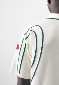 Knitted short-sleeve sweater in cream, featuring green striped details and a red label on the sleeve. Smooth texture with ribbed edges.