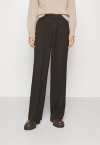 Pantalon taille haute, coupe large en tissu noir avec des plis sur le devant, doté de poches latérales et d'une texture lisse. Associé à des bottes marron épaisses.