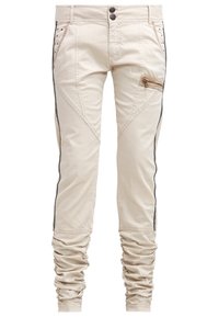 Jasnobeżowe spodnie o dopasowanym kroju, z bocznymi zamkami, czarnymi paskami po bokach, ozdobnym szyciem oraz marszczonymi mankietami przy kostkach.