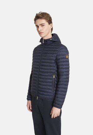 Jeune homme portant une veste matelassée à capuche bleu marine avec fermeture éclair à l'avant et poches latérales, debout devant un fond uni.