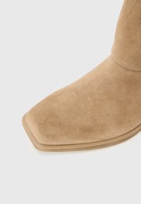 Bottine en suédine beige avec un bout carré et une semelle lisse. La tige présente une texture douce, mettant en avant le design minimaliste.