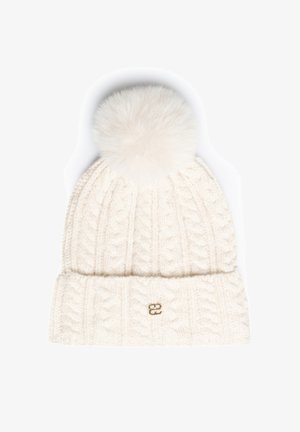 Balta megzta žieminė kepurė su pynių raštu, didelė pūsta bumbula viršuje ir mažu auksiniu logotipu ant sulenkto krašto.