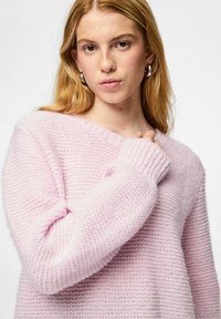 Pehmeä vaaleanpunainen neulepaita väljällä istuvuudella, V-kaula-aukoilla ja ribbineuloksella varustetuilla hihoilla. Siinä on kuvio, joka antaa lisälämpöä ja syvyyttä.