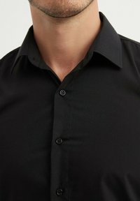 Chemise noire à boutons en tissu lisse, avec un col structuré, six boutons sur le devant et une coupe ajustée.
