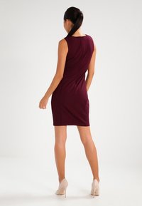 Sukienka bodycon w głębokim burgundzie, bez rękawów, do kolan, o dopasowanej sylwetce z subtelnymi detalami szwów, zestawiona z cielistymi szpilkami.