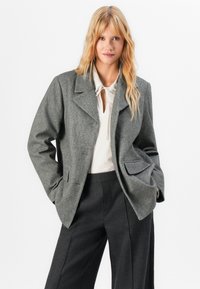 Blazer gessato grigio con colletto a punta, maniche lunghe e tasche frontali. Indossato sopra una blusa bianca e pantaloni neri.