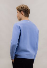 Lyseblå strikket genser med rund hals og ribbestrikkede mansjetter. Baksiden har en liten, diskret merkelapp nær halsen.