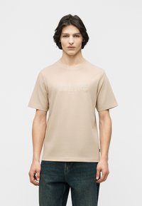 Νεαρός άνδρας με μεσαίου μήκους σκουρόχρωμα μαλλιά, ντυμένος με μπεζ κοντομάνικη μπλούζα με ανάγλυφο κείμενο  και μπλε τζιν, στέκεται μπροστά σε λευκό φόντο.