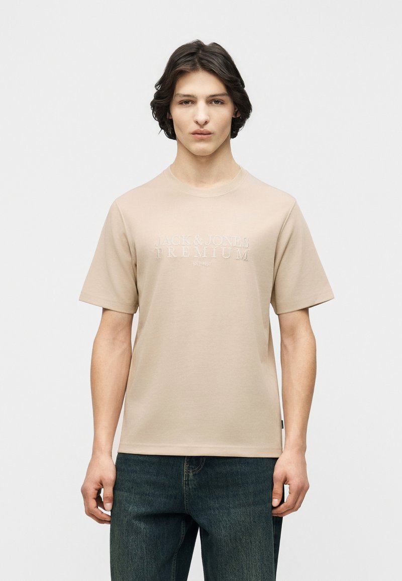 Νεαρός άνδρας με μεσαίου μήκους σκουρόχρωμα μαλλιά, ντυμένος με μπεζ κοντομάνικη μπλούζα με ανάγλυφο κείμενο και μπλε τζιν, στέκεται μπροστά σε λευκό φόντο.