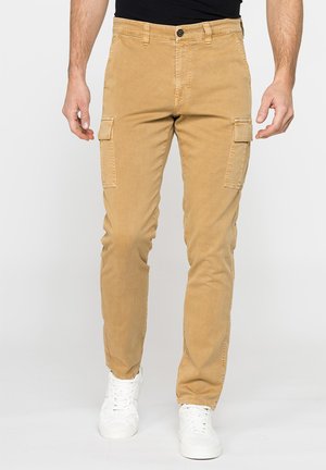 Uomo che indossa pantaloni cargo color sabbia con tasche a patta sui lati e sneakers bianche, in piedi davanti a uno sfondo chiaro e uniforme.