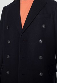Abrigo de mezcla de lana de doble botonadura en color azul marino con seis grandes botones negros, solapas con pico y un bolsillo en el pecho. Lleva una camisa naranja debajo.