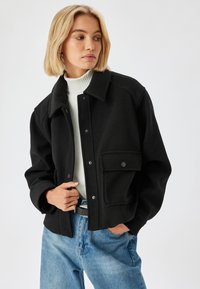 Schwarze, kurze Jacke mit Kragen, Knopfverschluss und Fronttasche. Aus strukturiertem Stoff gefertigt und kombiniert mit einem weißen Rollkragenpullover.