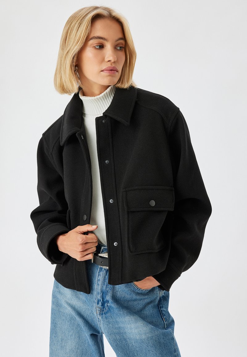 Schwarze, kurze Jacke mit Kragen, Knopfverschluss und Fronttasche. Aus strukturiertem Stoff gefertigt und kombiniert mit einem weißen Rollkragenpullover.