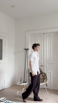 A modell fehér, rövid ujjú inget és fekete, bő szárú nadrágot visel, barna cipővel. A szobában egy állólámpa, egy kis asztal és egy szobor található.