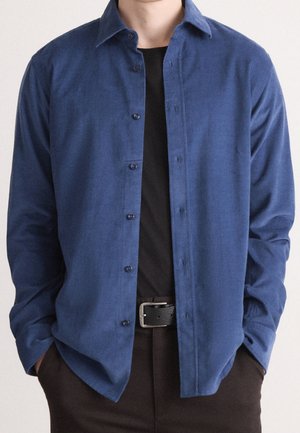 Man draagt een open blauwe lange mouwen overhemd over een zwart T-shirt met donker broek en een zwarte riem, handen in de zakken.