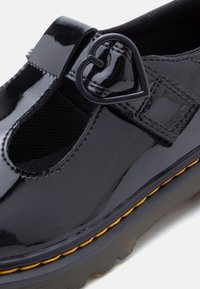 Chaussure en cuir verni noir avec un bout arrondi, des panneaux latéraux élastiques et une boucle noire en forme de cœur. Dispose d'une semelle texturée avec un accent jaune.