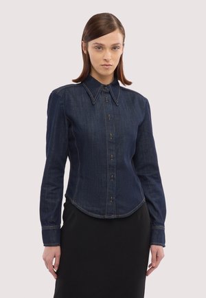 Kobieta ubrana w dopasowaną ciemną jeansową koszulę zapinaną na guziki z długimi rękawami i ostrym kołnierzykiem, w połączeniu z czarną spódnicą.