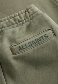 Pantaloni jogger verde oliva con un tessuto testurizzato, dotati di un'etichetta patch cucita che riporta la scritta "ALLSAINTS" in un font verde scuro.