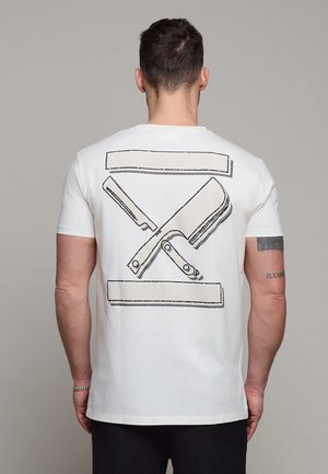 Uomo in piedi con la schiena rivolta verso la fotocamera che indossa una maglietta bianca con un'illustrazione nera contornata di un rasoio dritto e un tagliere incrociati sulla schiena.