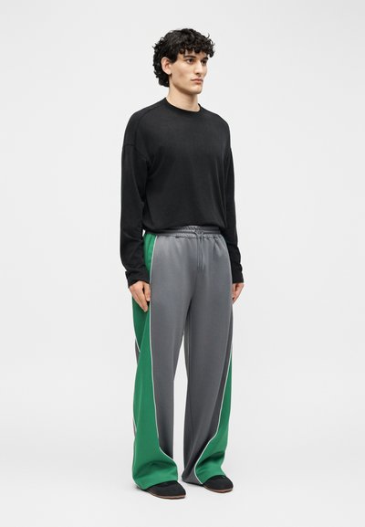 Άτομο με σγουρά μαλλιά, φορώντας μαύρο πουκάμισο με μακριά μανίκια και χαλαρό γκρι παντελόνι με πράσινες πλαϊνές πινελιές, στέκεται μπροστά σε απλό φόντο.