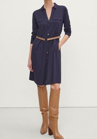 Robe chemise marine à manches longues, avec un devant à boutons, deux poches poitrine et une ceinture marron. Assortie à des bottes en cuir marron hautes.