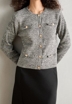 Woman wearing a gray textured knit cardigan with gold buttons and a black skirt, standing against a plain gray background.