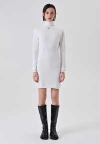 Giovane donna che indossa un vestito bianco a maniche lunghe con colletto alto e stivali neri sopra il ginocchio, in piedi contro uno sfondo chiaro e uniforme.