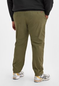 Olivgrüne Hose mit geradem Bein, ausgestattet mit Gesäßtaschen und einem dezenten Glanz, kombiniert mit sportlichen Schuhen mit metallischen Akzenten.