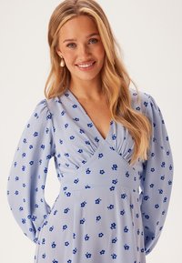 Lichtblauwe jurk met lange mouwen, V-hals en bloemmotief in donkerblauw. De stof is glad met een lichte glans en heeft een aansluitend lijfje.