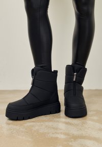 Černé zimní boty s krátkým siluetou mají prošívaný design, kulatou špičku a zip na vnější straně. Silné gumové podrážky zajišťují trakci.