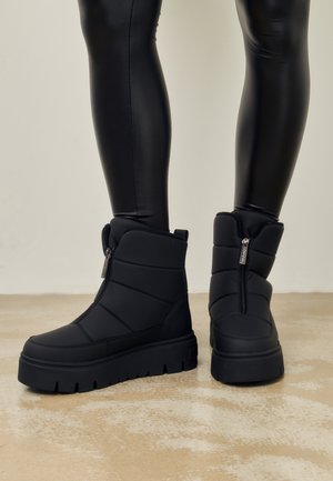 Zwarte pufferenkelschoenen hebben een doorstikte design, een ronde neus en een rits aan de buitenkant. Dikke rubberen zolen zorgen voor grip.
