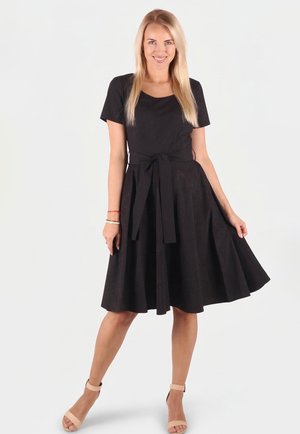 Blonde Frau lächelnd, trägt ein knielanges schwarzes Kleid mit gebundenem Taillengürtel, steht mit einer Hand am Rock, beige Knöchelriemen-Pumps.