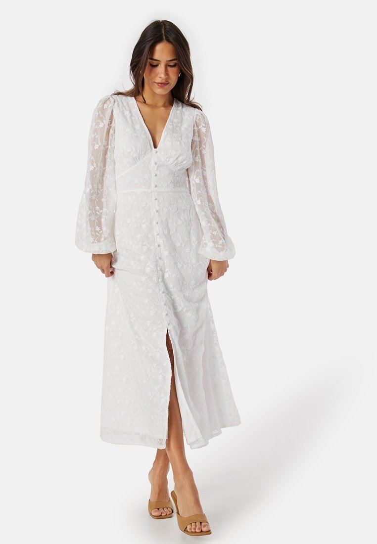 Vit maxiklänning med blommönsterbroderi, genomskinliga långärmar, V-ringning, framknäppning och en hög slits fram. Bärs med beige klackar.