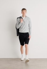 Šedá mikina na zip, černé šortky a bílé tenisky. Model drží šedou bundu přes rameno na jednobarevném pozadí.