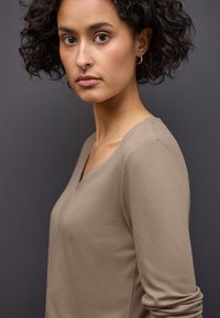 Mladá žena s kudrnatými tmavými vlasy a zlatými kruhovými náušnicemi, oblečená v béžovém topu s V výstřihem, obrácená mírně na stranu na tmavě šedém pozadí.