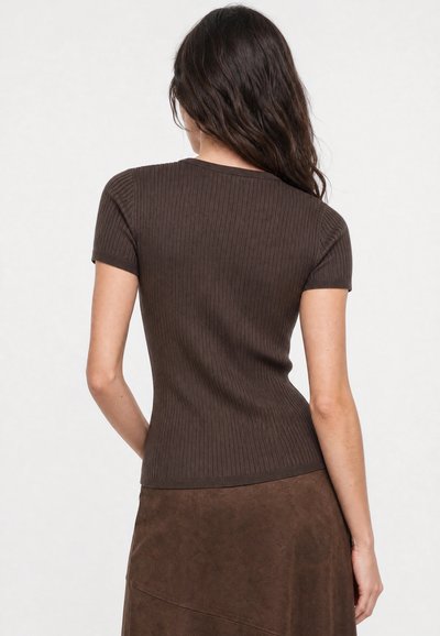 Femme aux longs cheveux foncés porte un haut marron côtelé à manches courtes et ajusté ainsi qu'une jupe marron, tournée de dos sur un fond uni.