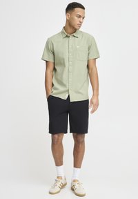 Short-sleeve green textured shirt with a pocket, paired with black shorts. Model wears white socks and beige athletic shoes.