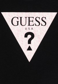 Melna T-krekls ar rozā trīsstūra grafiku, kas satur teksturētu rakstu, uzrakstu "GUESS" treknā fontā un jautājuma zīmi centrā.